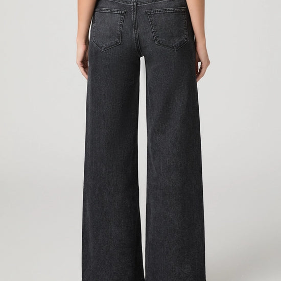 Rear view of a model wearing dark gray wide-leg denim jeans with a faded wash, paired with a dark red top and black heels, against a light gray studio background.