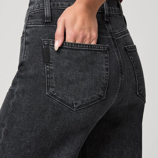 Close-up back view of a person wearing dark charcoal gray washed jeans, with one hand tucked into the back pocket, against a light gray studio background.
