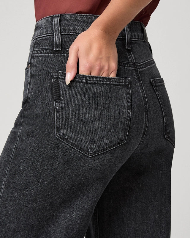 Close-up back view of a person wearing dark charcoal gray washed jeans, with one hand tucked into the back pocket, against a light gray studio background.