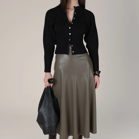 A model stands against a light beige studio background wearing a black button-front cardigan, an olive green faux-leather midi skirt, dark brown pointed-toe boots, and holding a black leather tote bag.