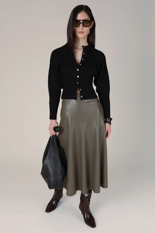 A model stands against a light beige studio background wearing a black button-front cardigan, an olive green faux-leather midi skirt, dark brown pointed-toe boots, and holding a black leather tote bag.