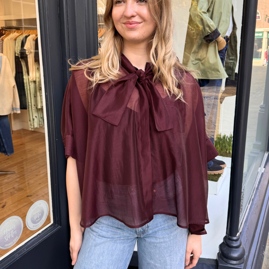 Women wearing burgundy sheer blouse with tie neck and blue jeans.