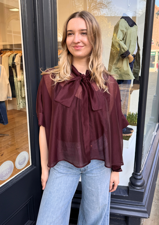 Women wearing burgundy sheer blouse with tie neck and blue jeans.