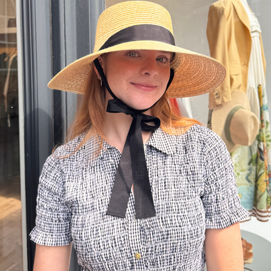 Woman wearing a wide-brimmed hat with a black ribbon in front of a store window.