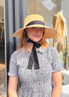 Woman wearing a wide-brimmed hat with a black ribbon in front of a store window.