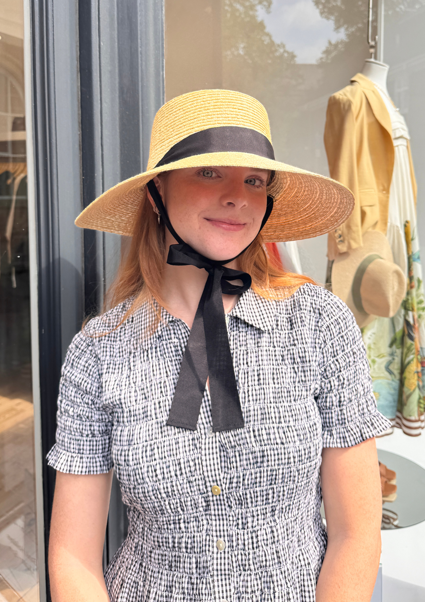 Woman wearing a wide-brimmed hat with a black ribbon in front of a store window.
