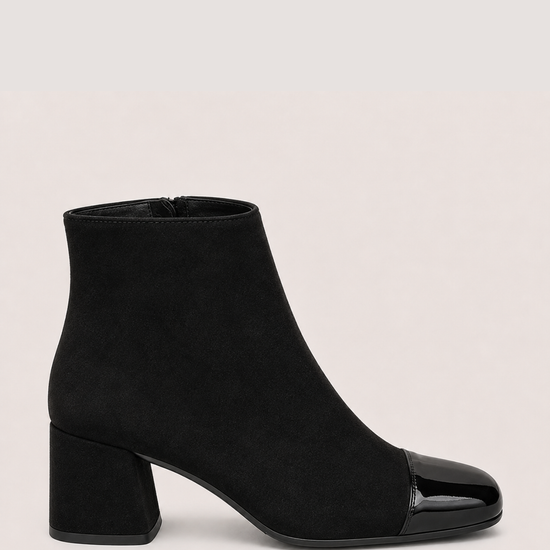 A black ankle boot with a matte suede-like body and a glossy patent leather toe cap, set against a plain light beige background.