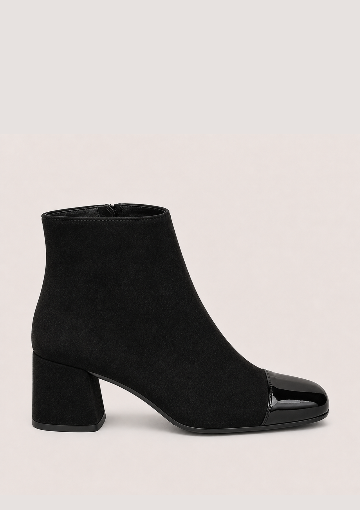 A black ankle boot with a matte suede-like body and a glossy patent leather toe cap, set against a plain light beige background.