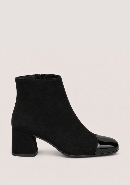A black ankle boot with a matte suede-like body and a glossy patent leather toe cap, set against a plain light beige background.