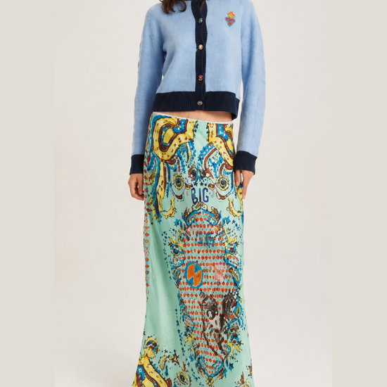 A model wearing a light blue cropped cardigan with dark navy trim and a long, colorful patterned skirt in shades of mint green, yellow, and blue, paired with dark brown ankle boots, against a light gray studio background.
