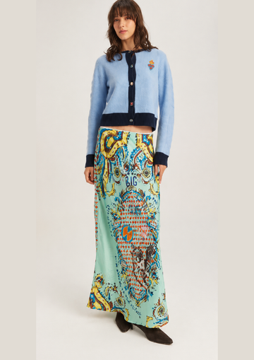 A model wearing a light blue cropped cardigan with dark navy trim and a long, colorful patterned skirt in shades of mint green, yellow, and blue, paired with dark brown ankle boots, against a light gray studio background.
