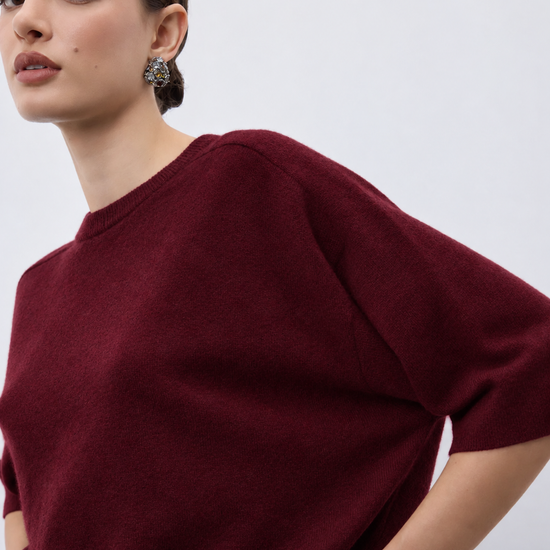 A close-up of a model wearing a short-sleeve crewneck knit sweater in deep burgundy, paired with a gold chain bracelet and a blue-green gemstone earring, against a plain light gray background.