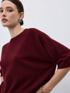 A close-up of a model wearing a short-sleeve crewneck knit sweater in deep burgundy, paired with a gold chain bracelet and a blue-green gemstone earring, against a plain light gray background.