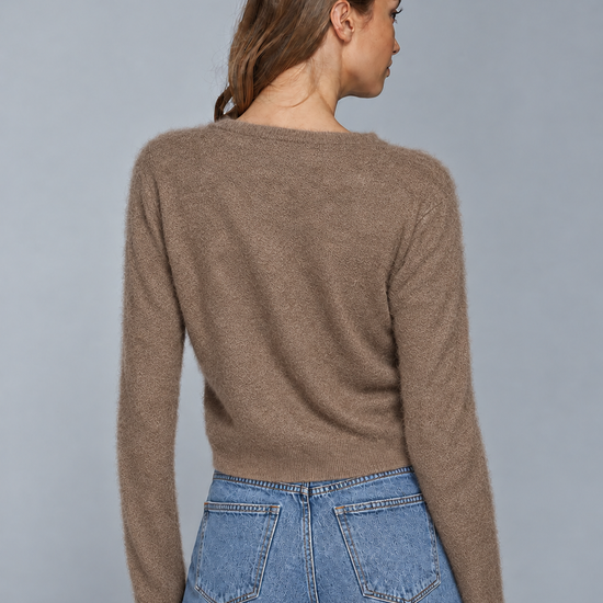 A woman shown from the back wearing a cropped, long-sleeve knit sweater in a warm taupe-brown color, paired with light-wash blue jeans, against a solid gray studio background.