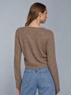 A woman shown from the back wearing a cropped, long-sleeve knit sweater in a warm taupe-brown color, paired with light-wash blue jeans, against a solid gray studio background.