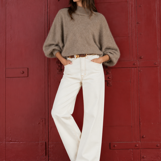 A woman stands in front of a large red metal door wearing a taupe-brown oversized knit turtleneck sweater, white wide-leg pants, and dark brown pointed-toe flats.