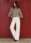 A woman stands in front of a large red metal door wearing a taupe-brown oversized knit turtleneck sweater, white wide-leg pants, and dark brown pointed-toe flats.