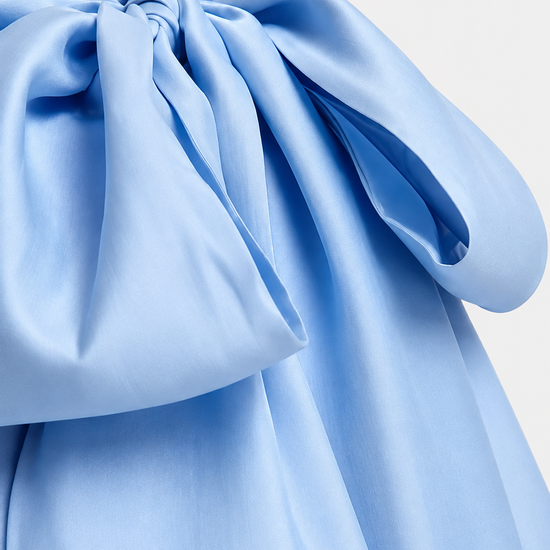 A close-up of a light blue satin-like fabric with a large gathered bow or tie detail, showing soft folds and sheen against a plain light background.