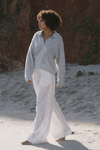 Woman in light blue top and white pants standing on a sandy beach with a blurred natural background