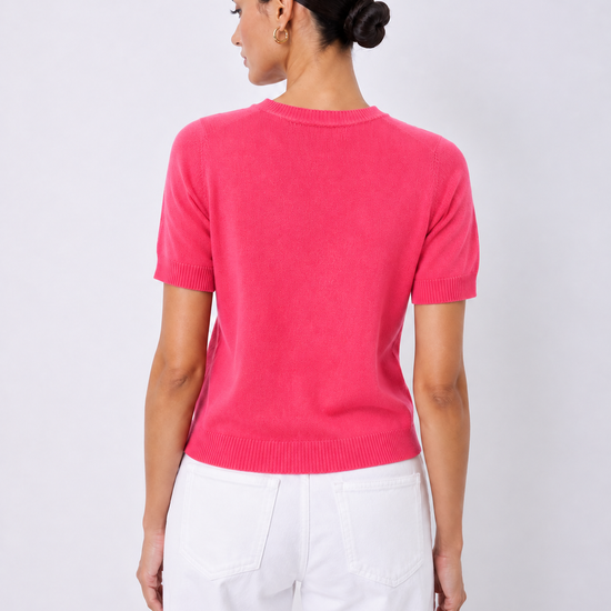 A woman shown from the back wearing a bright pink short-sleeve knit sweater and white pants, standing against a light gray studio background.