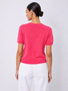 A woman shown from the back wearing a bright pink short-sleeve knit sweater and white pants, standing against a light gray studio background.