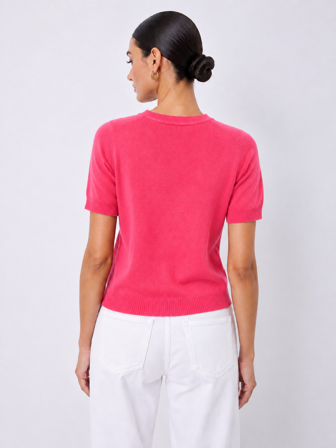 A woman shown from the back wearing a bright pink short-sleeve knit sweater and white pants, standing against a light gray studio background.