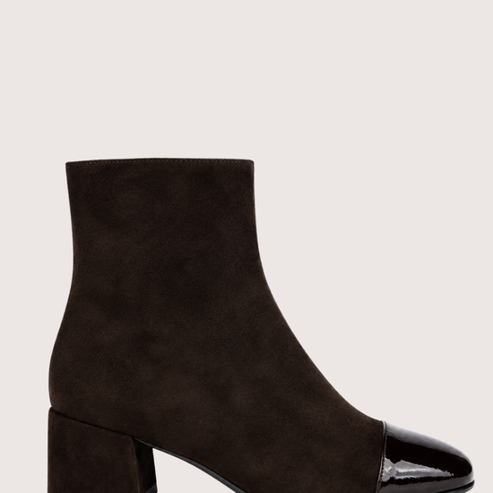 A pair of dark brown suede ankle boots with glossy patent leather cap toes and low block heels, shown against a light beige background.
