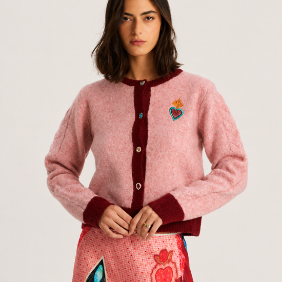 A model wearing a pink fuzzy knit cardigan with dark red ribbed trim and a colorful embroidered heart patch on the chest, paired with a patterned skirt, against a light gray studio background.