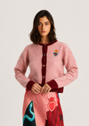 A model wearing a pink fuzzy knit cardigan with dark red ribbed trim and a colorful embroidered heart patch on the chest, paired with a patterned skirt, against a light gray studio background.