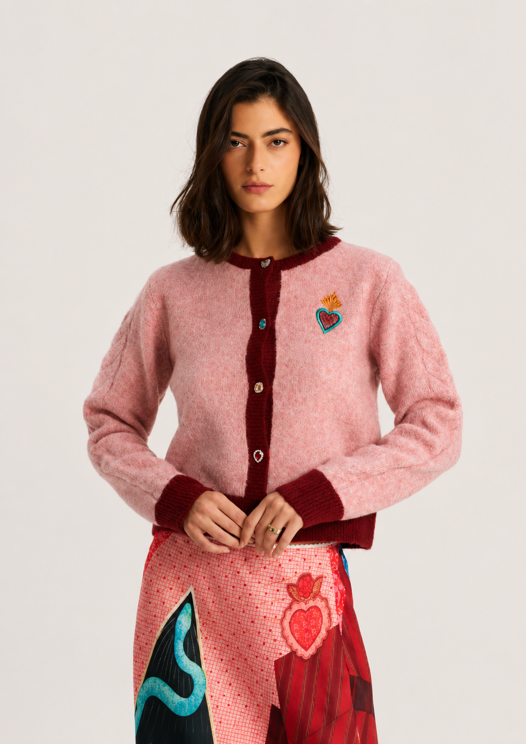 A model wearing a pink fuzzy knit cardigan with dark red ribbed trim and a colorful embroidered heart patch on the chest, paired with a patterned skirt, against a light gray studio background.