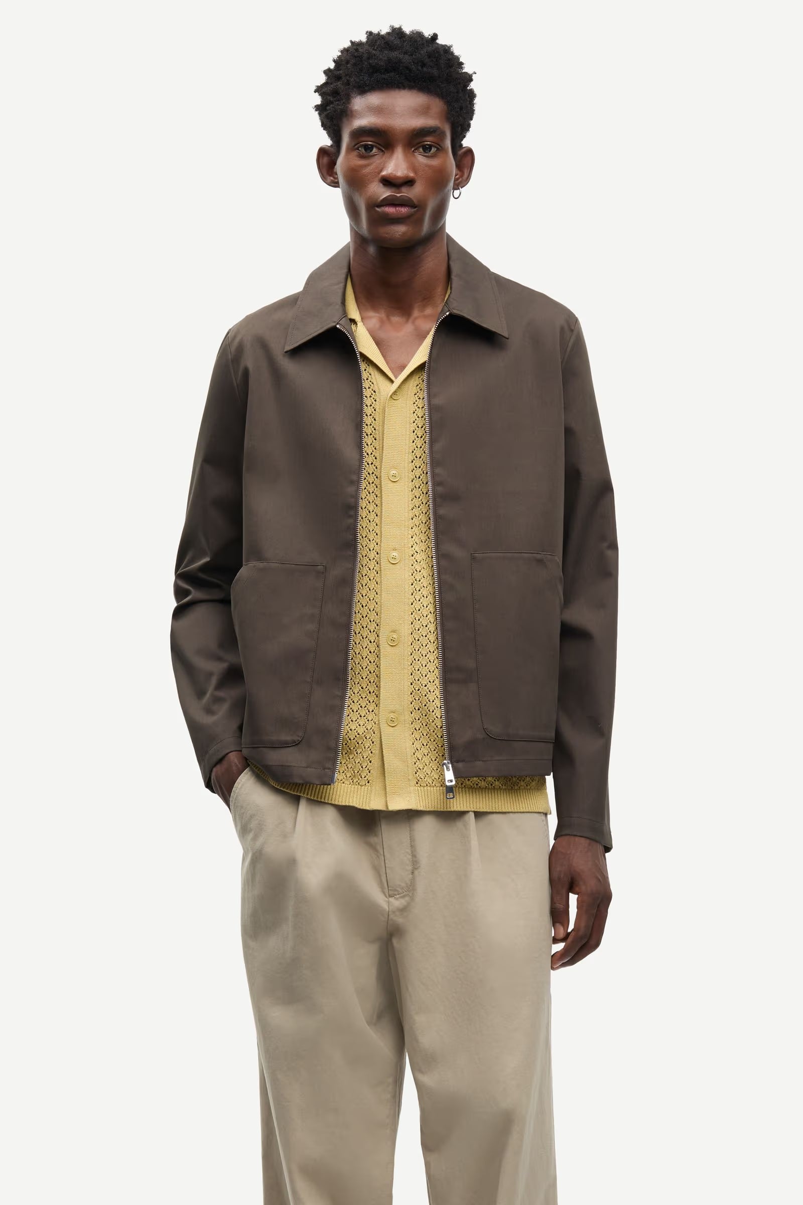 Man wearing a brown jacket by Samsoe Samsoe over a yellow shirt with beige pants on a white background