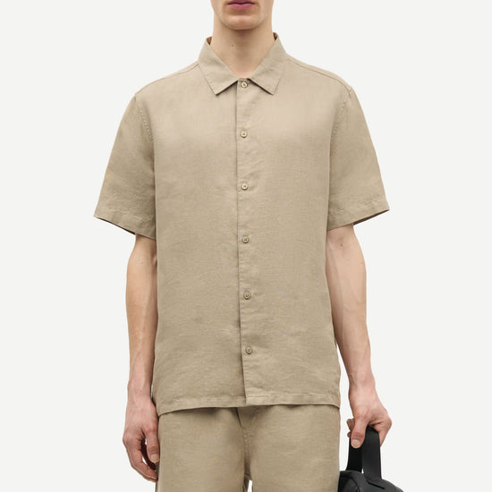 Man wearing a beige shirt and shorts by Samsoe Samsoe holding a black backpack on a white background