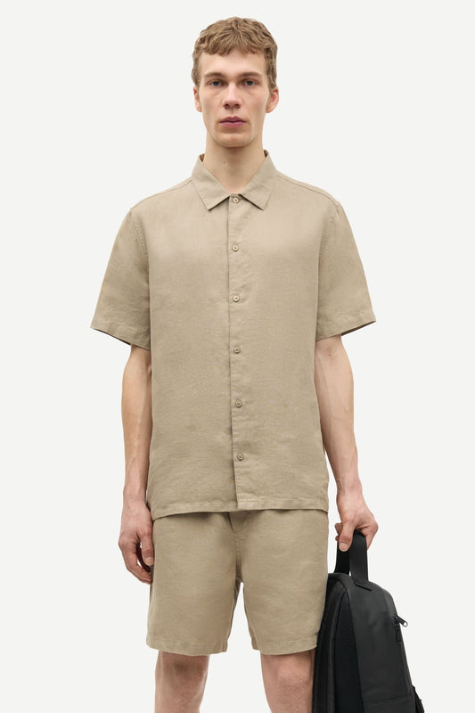 Man wearing a beige shirt and shorts by Samsoe Samsoe holding a black backpack on a white background