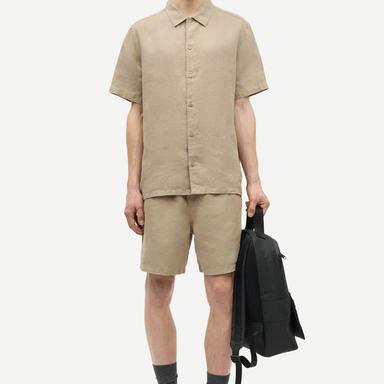 Man wearing a beige shirt and shorts by Samsoe Samsoe holding a black backpack on a white background
