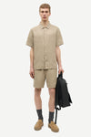 Man wearing a beige shirt and shorts by Samsoe Samsoe holding a black backpack on a white background
