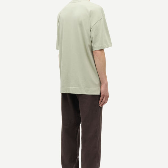 Man wearing a light green t-shirt and brown pants by Samsoe Samsoe on a white background rear view