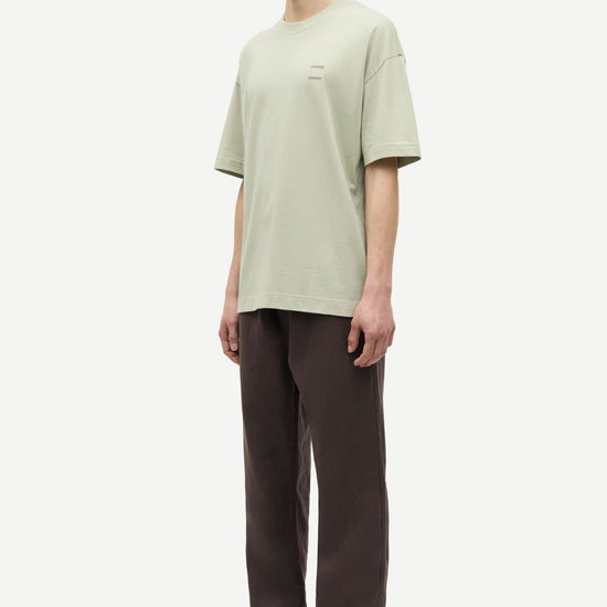 Man wearing a light green t-shirt and brown pants by Samsoe Samsoe on a white background side view