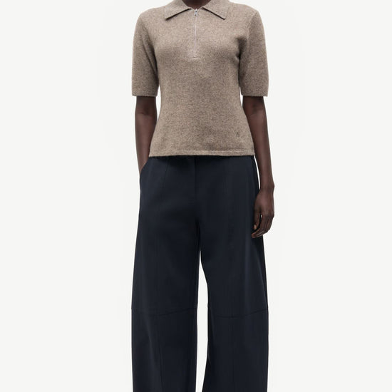 A model stands in profile wearing a short-sleeve knit polo shirt in a light taupe-beige color and wide-leg dark navy trousers, photographed against a clean off-white studio background.