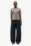 A model stands in profile wearing a short-sleeve knit polo shirt in a light taupe-beige color and wide-leg dark navy trousers, photographed against a clean off-white studio background.