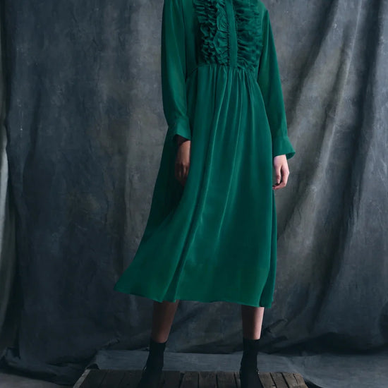 Woman wearing a green dress standing on a wooden platform against a dark gray curtain.
