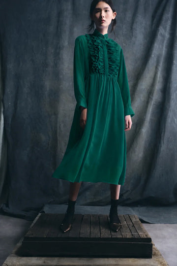 Woman wearing a green dress standing on a wooden platform against a dark gray curtain.
