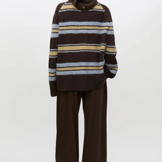 A model viewed from the back wearing a brown knit sweater with horizontal stripes in beige, light blue, and white, paired with wide-leg brown trousers, against a plain white studio background.