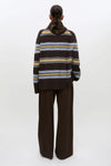 A model viewed from the back wearing a brown knit sweater with horizontal stripes in beige, light blue, and white, paired with wide-leg brown trousers, against a plain white studio background.