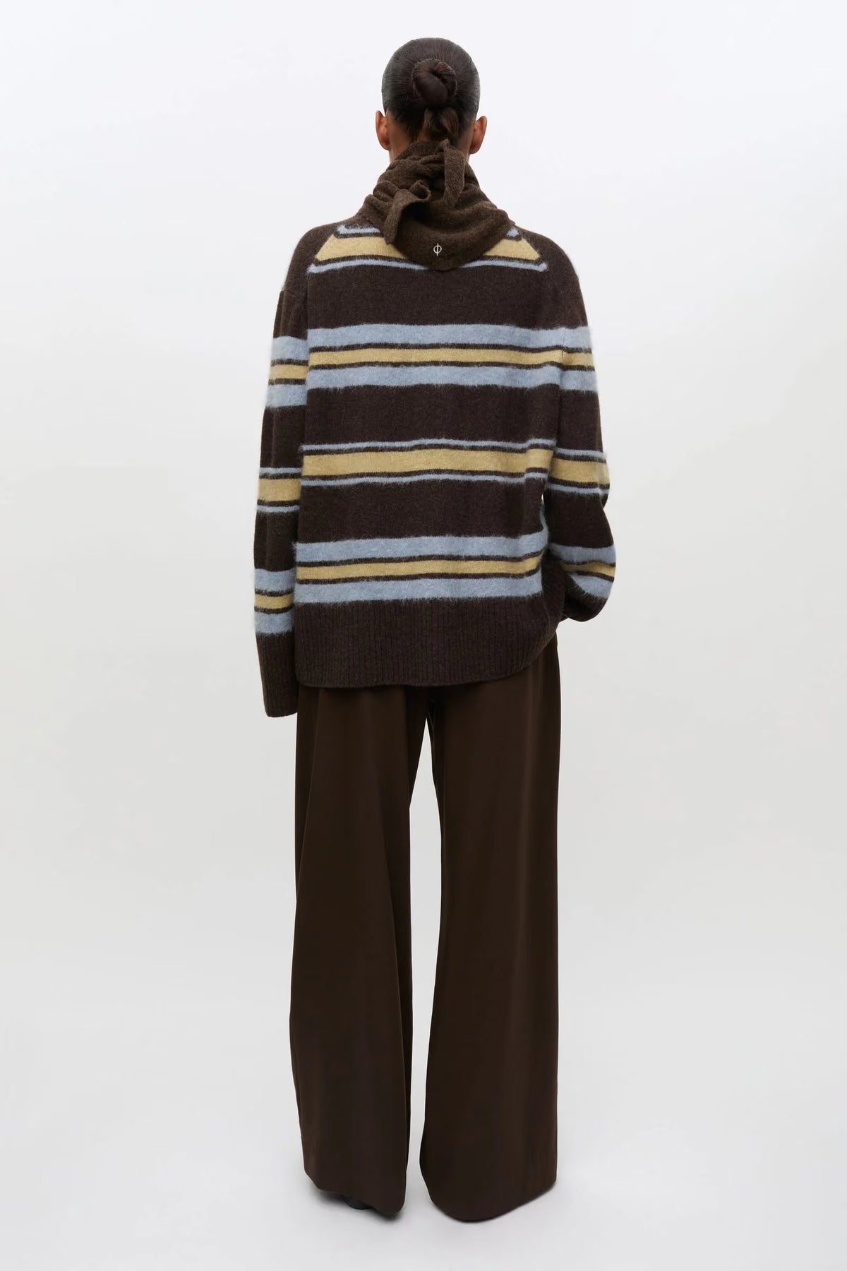 A model viewed from the back wearing a brown knit sweater with horizontal stripes in beige, light blue, and white, paired with wide-leg brown trousers, against a plain white studio background.