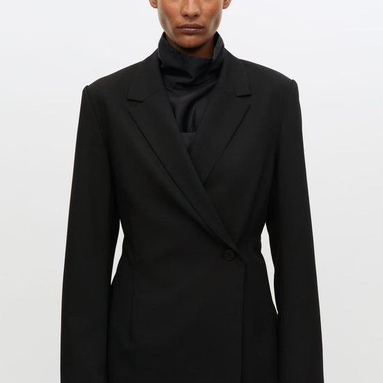 A model wearing a black structured double-breasted blazer over a black ruffled high-neck top, photographed against a plain light gray studio background.