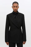 A model wearing a black structured double-breasted blazer over a black ruffled high-neck top, photographed against a plain light gray studio background.
