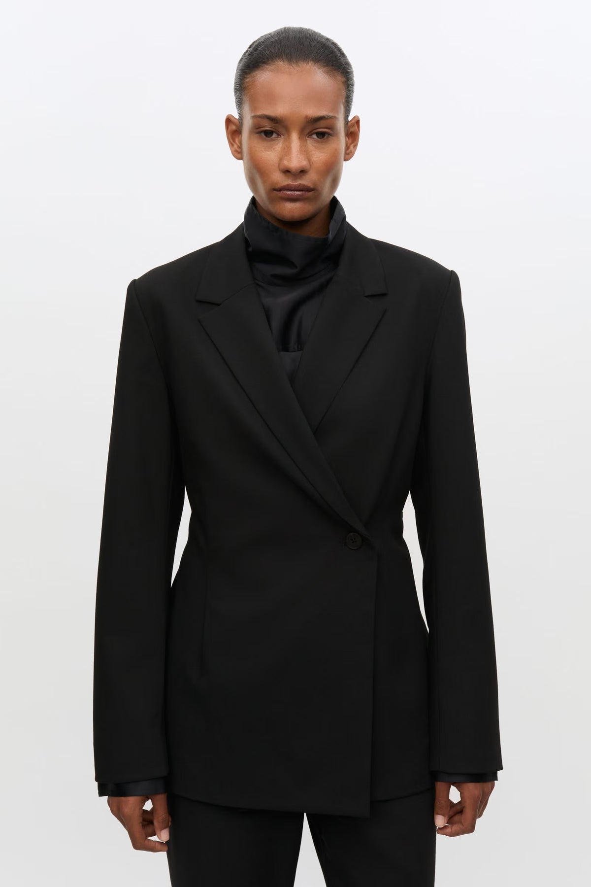 A model wearing a black structured double-breasted blazer over a black ruffled high-neck top, photographed against a plain light gray studio background.
