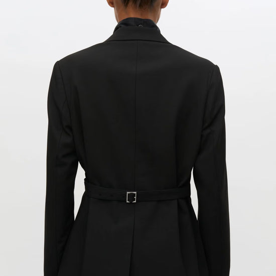 A person shown from the back wearing a black belted blazer with a high collar, photographed against a plain light gray studio background.