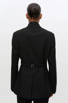 A person shown from the back wearing a black belted blazer with a high collar, photographed against a plain light gray studio background.