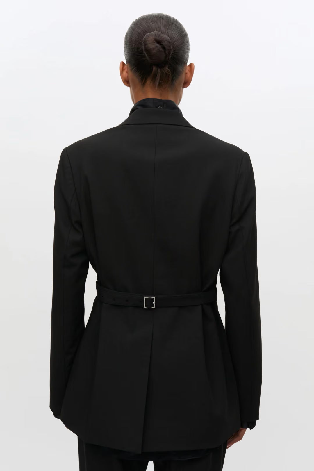 A person shown from the back wearing a black belted blazer with a high collar, photographed against a plain light gray studio background.